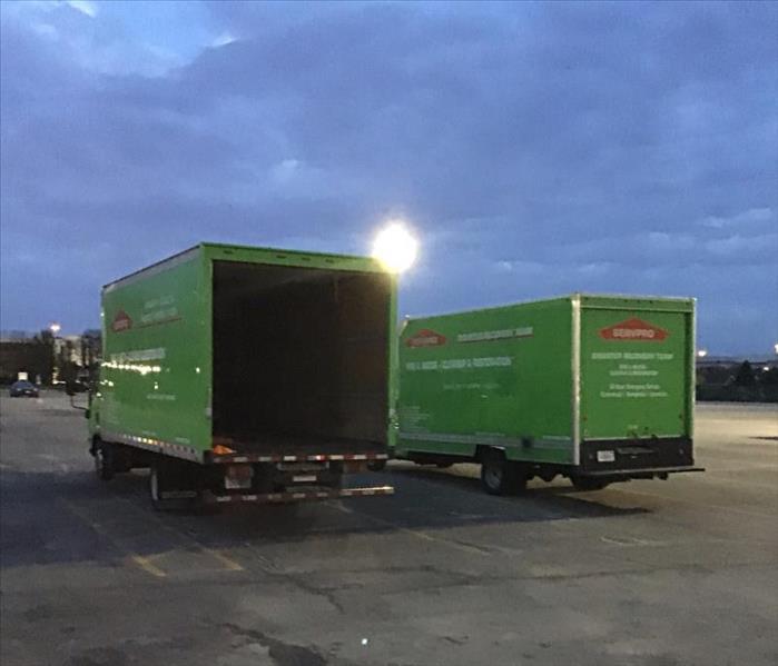 Sun setting over two big green SERVPRO box trucks that are parked in a parking lot.